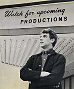 Photograph of Stan Klees in front of a sign reading, WATCH FOR UPCOMING PRODUCTIONS