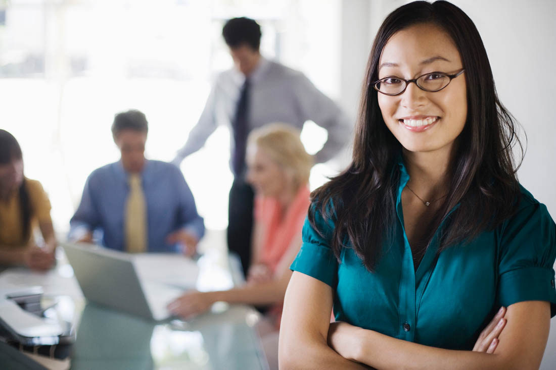 Photo – Dans un bureau, une femme sourit à la caméra pendant qu’un groupe d’individus ont une réunion dans l’arrière-plan.