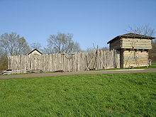 A wooden stockade fronted by a wall of vertically-placed logs and a corner blockhouse