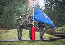 Three soldiers in camouflage stand in salute while a forth raises a blue and white flag on a red and white stripped flagpole.