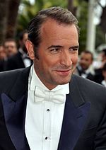 A black-haired man wearing a bowtie and a white shirt over a black suit is smiling. Several men are seen behind him in the distance.