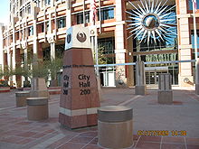 photos showing the short obelisk signage showing City Hall, and topped with the seal of the city, a stylized maroon phoenix.  The semi-circular front of the building in the background, adorned with a stylized sunburst.