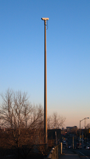 A video camera mounted on a tall cement pole on the side of a roadway. The camera is not pointing at the roadway visible at the bottom-right of the picture, but to the left.