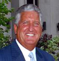 A portrait of a tan man with white-gray hair in a blue suit, smiling.