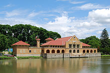 An orange-red mission-style building is seen on the banks of a lake, surrounded by trees.