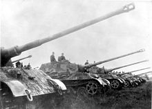 A row of seven large German tanks from World War Two lined up with their long cannons pointing up at an angle, as if saluting.