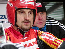 A man with a goatee wears a red-white-and-yellow tight jumpsuit, with a red-and-white vest over it, and a red helmet with a raised full-faced visor. He shows a concentrated look. Partially hidden behind him is another man, wearing a black tracksuit jacket and winter cap.