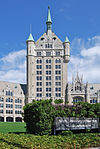 An ornate stone tower with a pointed green roof flanked by similar small turrets, and similar but lower wings at ground level. At front right is a shrub with a sign in front of it saying "State University of New York, System Administration