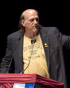 Balding man with long hair, T-shirt and jacket at podium.