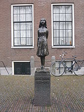 A bronze statue of a smiling Anne Frank, wearing a short dress and standing with her arms behind her back, sits upon a stone plinth with a plaque reading "Anne Frank 1929–1945". The statue is in a small square, and behind it is a brick building with two large windows, and a bicycle. The statue stands between the two windows.