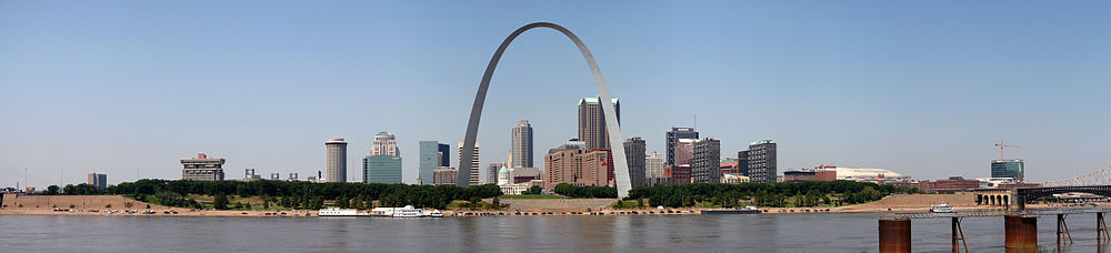 A large arch is in the center, across from a river. A clump of tall buildings is scattered behind it.