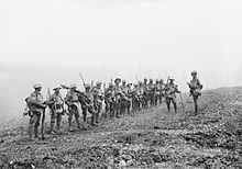 A line of soldiers in battle equipment face another soldier who is addressing them on a gentle slope.  Behind them, smoke or fog obscures the rest of the terrain