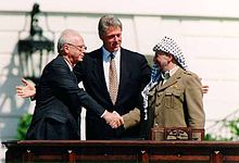 A man in a dark suit on the left shakes the hand of a man in traditional Arab headdress on the right. Another man stands with open arms in the center behind them.