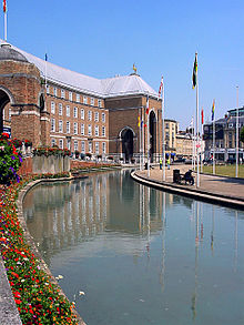 A large brick building, built in a shallow curve, with a central porch. In front of that a pool and a water fountain.