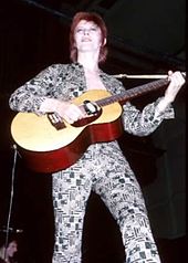 A color photograph of David Bowie with an acoustic guitar