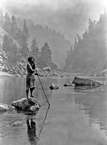 Photo of man standing on rock holding spear with spearpoint in the water