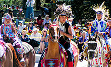 "Colour photograph of Tsuu T'ina children in traditional costume on horseback at a Stampede Parade in front of an audience"