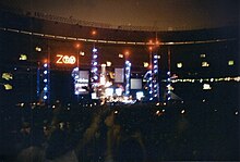 An elaborate concert stage set bearing a logo that reads "Zoo TV", set in a dark stadium. Towers reach into the night sky, illuminated in blue with red warning lights on top.