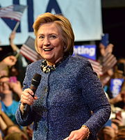 Hillary Clinton, a 68-year-old white woman with blonde hair, holding a microphone and wearing a blue suit.