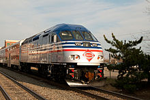 Blue and silver train engine with red and white accent lines moves closer leading a series of similarly colored passenger cars with shrubs and a sound wall in the background.