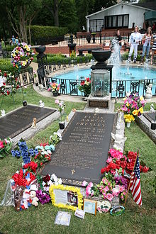 A long, ground-level gravestone reads "Elvis Aaron Presley", followed by the singer's dates, the names of his parents and daughter, and several paragraphs of smaller text. It is surrounded by flowers, a small American flag, and other offerings. Similar grave markers are visible on either side. In the background is a small round pool, with a low decorative metal fence and several fountains.