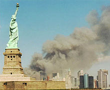 A monumental green copper statue of a woman with a torch stands on an island in front of a mainland where a massive plume of gray smoke billows amongst skyscrapers.
