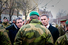 Two gray haired older men talk with a soldier wearing camouflage and a green beret who is facing away.