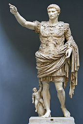 A marble statue of the Emperor Augustus. He stands with one arm raised as if in command. Augustus is depicted as a man of about thirty five, with short hair and clean shaven. He wears Roman military uniform of a breast plate, leather accoutrements and a cloak over a short tunic. The breastplate is decorated with symbolic figures. As a work of art, the statue displays high technical mastery.