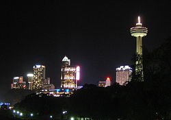A spike tower with a pod is lit along its length on the right. Other buildings show interior lights in this night scene. The foreground is a deep black sky.