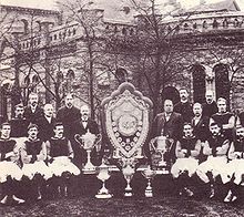 A sepia photograph with a large old structure in the background obscured by trees. In the foreground there is a large shield surrounded by five trophies. On either side of the shield stands 8 people.