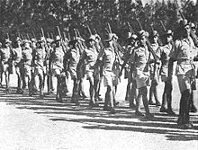 Soldiers in colonial-era military uniform march past, rifles shouldered.