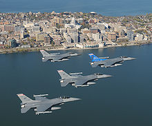  Four jets flying right in formation over water. In the foreground are buildings erected on a narrow piece of land, with water on both sides