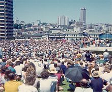 A large crowd has gathered in an open space in an urban setting (tall buildings are visible).  Far away can be seen a platform, and a banner reading SHAME FRASER SHAME.
