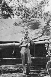 Whitlam in military uniform stands under a tree in front of a large tent. He holds a mug in his hand.