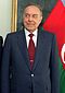 A man in a dark suit with a red tie standing in front of the Azerbaijani flag