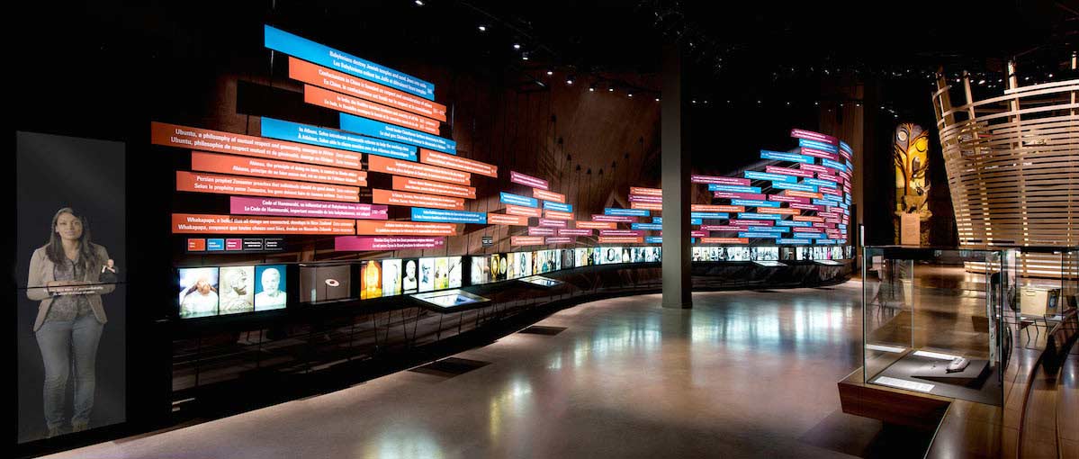 Panels with text are displayed in a large gallery. On the left, a woman is on a screen. A box made of glass is on the right.