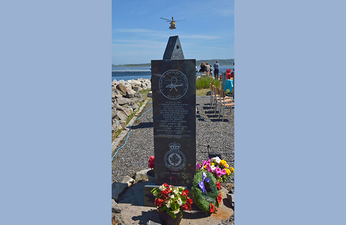 slide - A narrow, black granite, engraved memorial stone is in the foreground as a yellow Cormorant helicopter approaches in the distance.