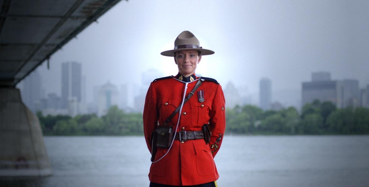 Image of a Royal Canadian Mounted Police officer in dress uniform standing at ease with a city skyline in the background. 