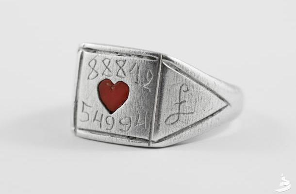 A close-up of a silver ring with a red heart shape in its centre. The ring also has two numbers carved into it, above and below the heat.  Above, the number is 88842 and below, the number is 54994.  A letter “L” is also visible on the side of the ring.