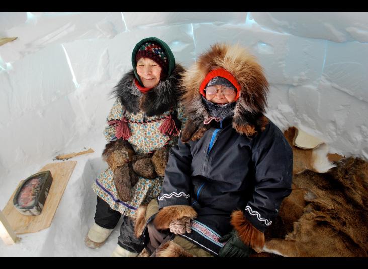 Two women in winter coats sit inside an igloo.