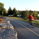 A biker rushing along the bike path that follows the Lairet River in Cartier-Brébeuf National Historic Site, with a view of the river and the city in the background.