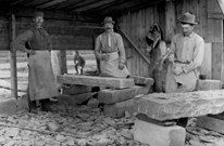 Four men stand next to the stones they're carving in a workshed. Archive #PA130013.