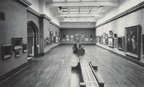 A large room with benches in the centre and paintings hanging on the walls. Archive #PA42658N.