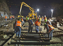 A crew of men in orange vests with reflector stripes and neon green hard hats over their clothing laying new railroad ties. Behind them is a large yellow piece of heavy equipment. It is night, and the scene is illuminated by large lights.