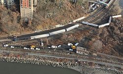 An aerial view of a wooded area between two watery areas in late autumn, with two railroad tracks forking to the top near the left and one following the water's edge. Several silvery train cars and a locomotive are lying on their sides off the tracks to the top; there are many parked yellow and white trucks and other vehicles between the two tracks.