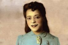 A head-and-shoulder portrait of a smiling Viola Desmond. She is wearing a light blue jacket with embroidered patterns on it. The jacket is held together at the collar by a large pin in the shape of a hand making a “V for victory” symbol.