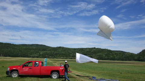 Un ballon blanc et un filet à papillons flottent dans le ciel, tous deux retenus au sol par des cordes attachées à un camion.