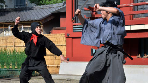 Deux hommes ninjas participent à un combat face à face à l'extérieur, celui de droite tenant un sabre à la main. 