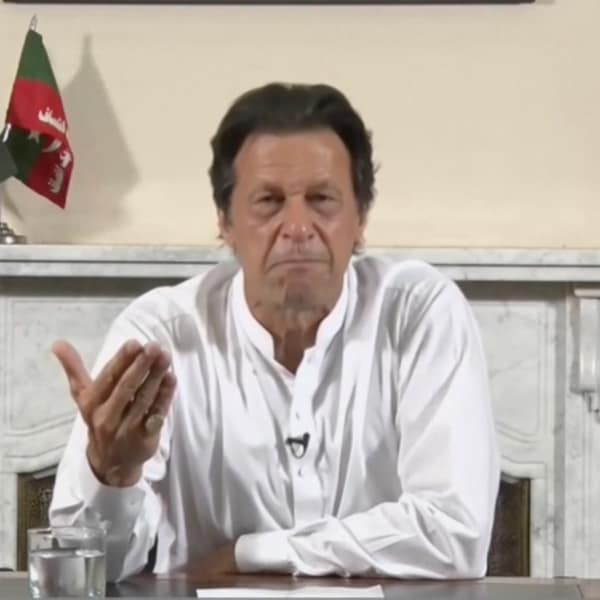 Un homme en chemise, assis à une table, s'adresse à la caméra. Derrière lui des drapeaux de son parti et du Pakistan.