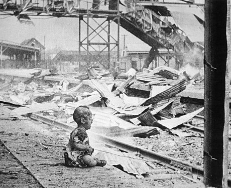 Black-and-white photograph of a baby crying on a platform of Shanghai's South Station after aerial bombing by Japanese forces, their clothes burned and their skin sooty, with flaming wreckage in the background